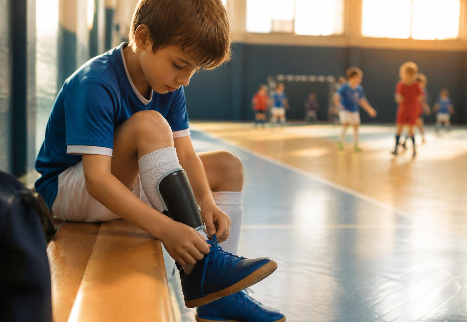 Menino sentado no banco de uma quadra de futsal colocando a chuteira, com outras crianças jogando ao fundo